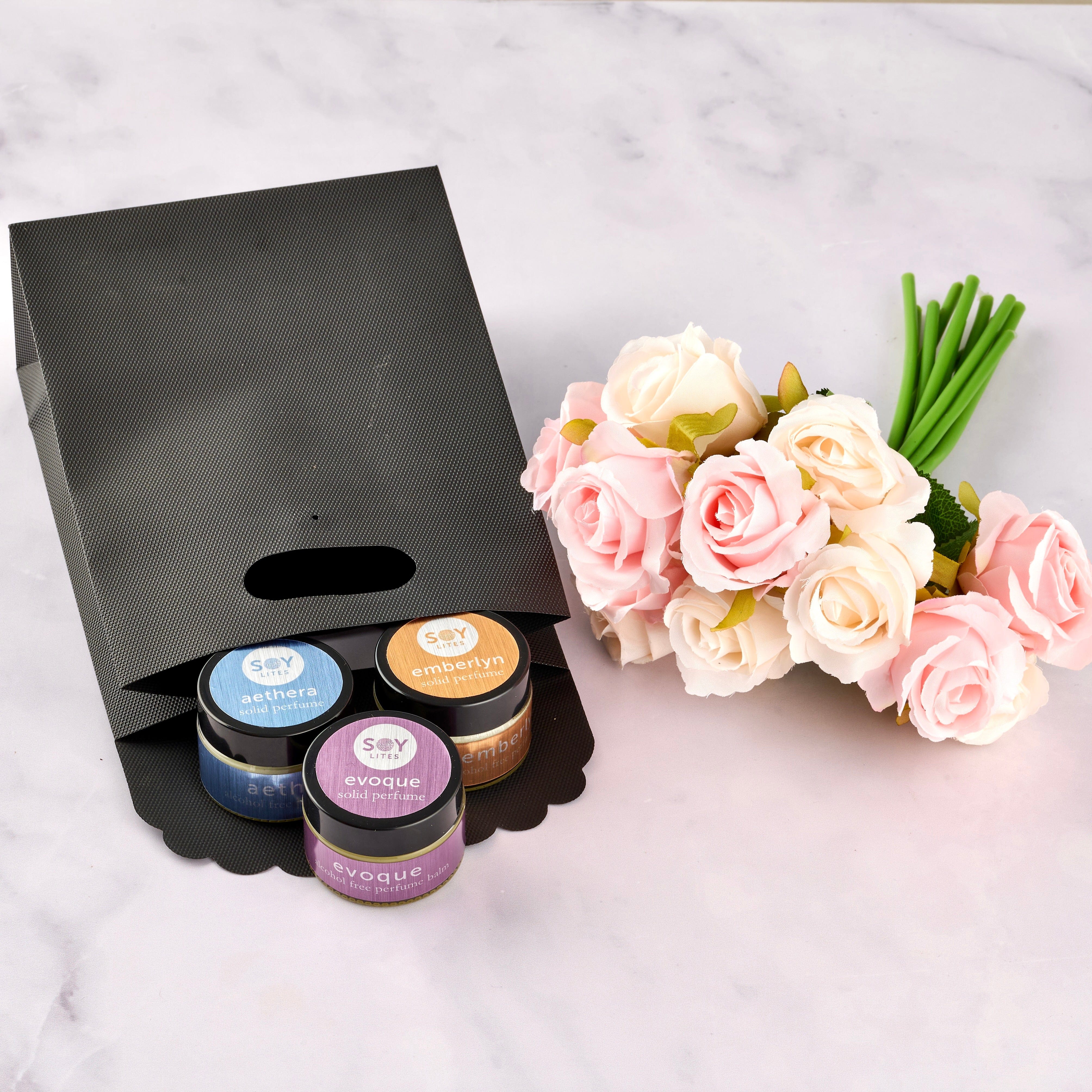 Set of small jars of soy solid perfumes with visible labels next to a bouquet of flowers on a marble surface