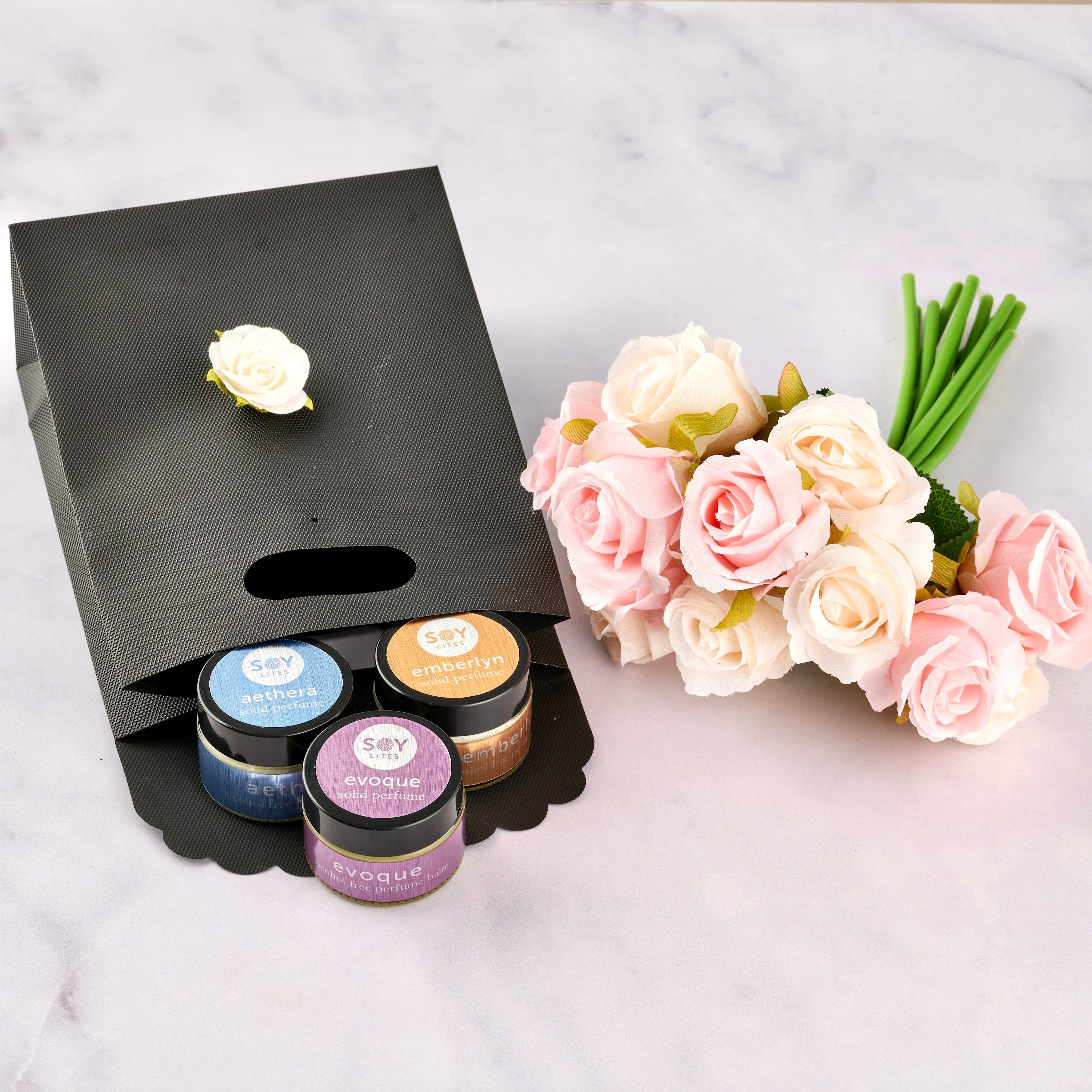 Set of small jars with lids of solid perfumes, a black stand with flowers, and a bouquet of pink and white roses on a light grey background.